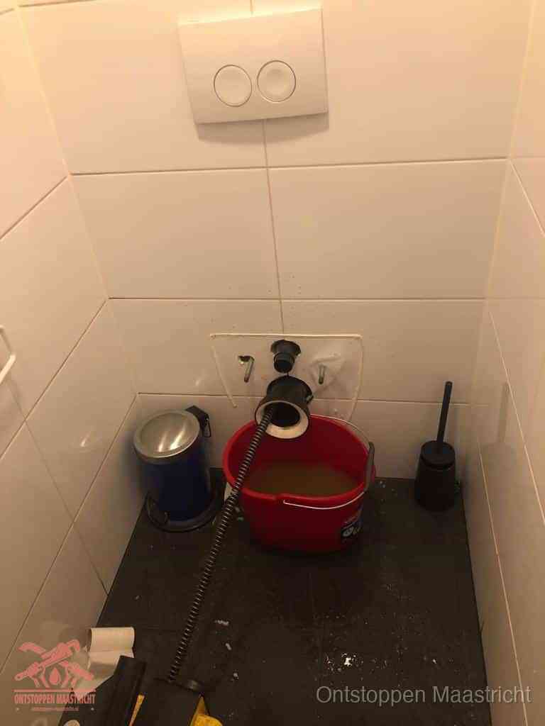 Een ontstopte wand-wc met verwijderde toiletpot waar een ontstoppingsveer in de wanduitgang verdwijnt, het achterhalen waar de verstopping zit in Maastricht
