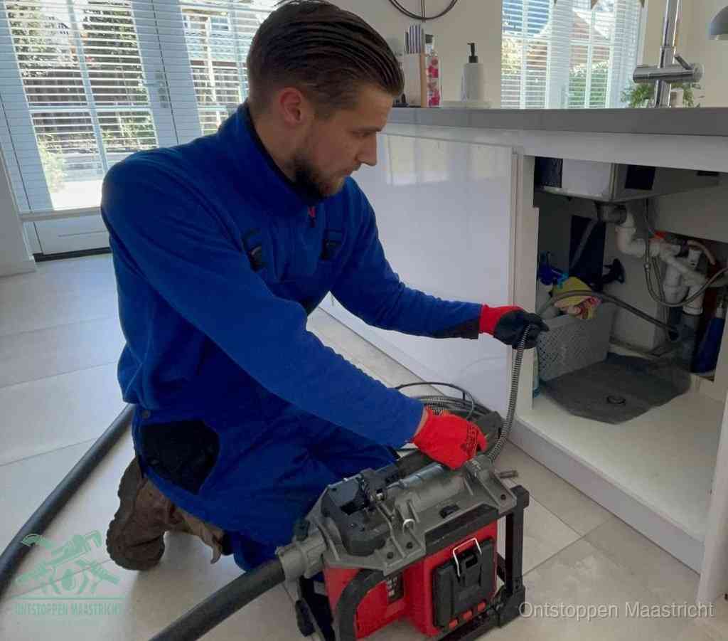 Een monteur met rode handschoenen knielt in een keuken en voert een elektrische ontstoppingsveer in tijdens het professioneel afvoer ontstoppen in Maastricht